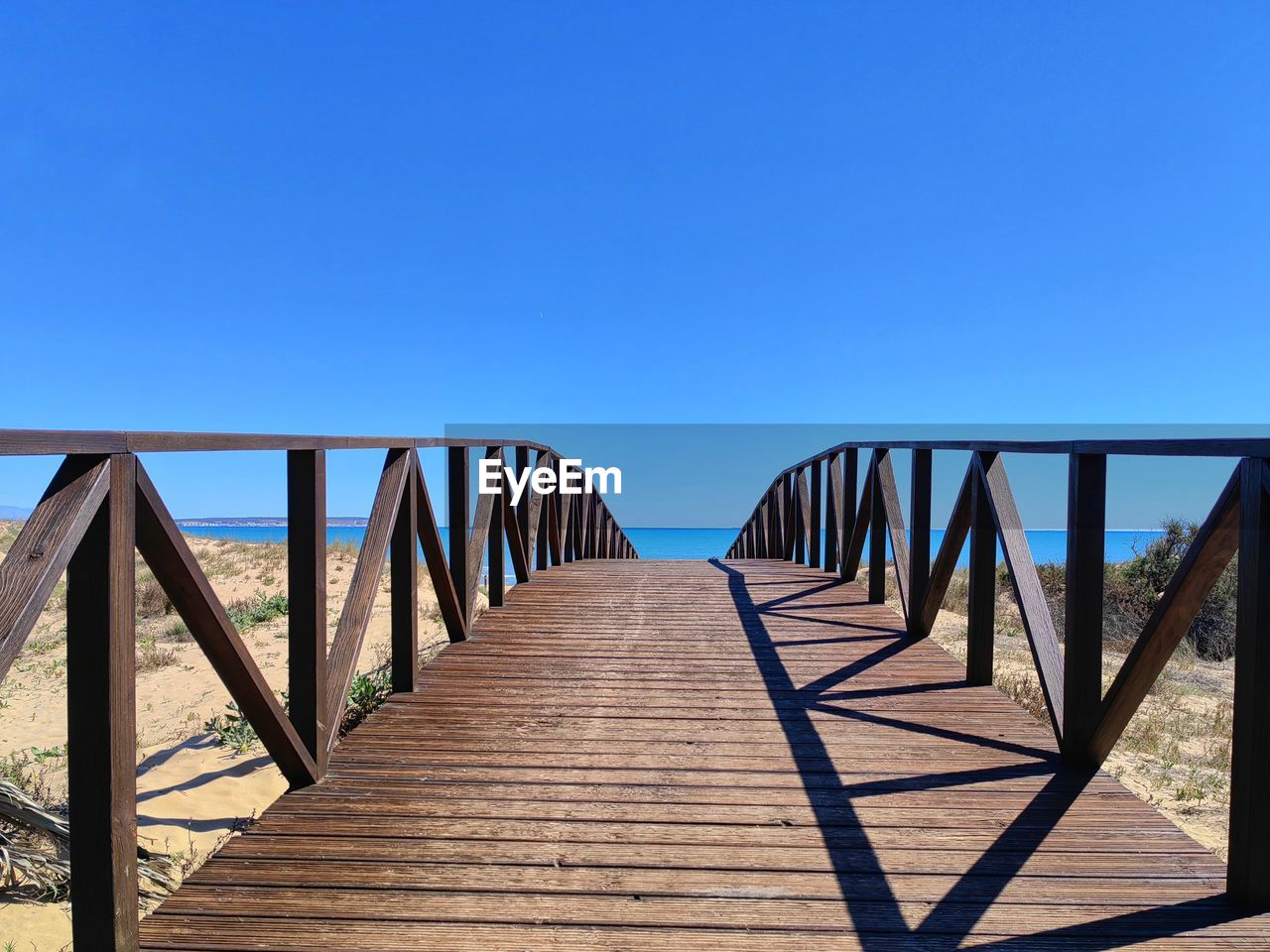 sky, wood, blue, architecture, nature, clear sky, walkway, built structure, bridge, water, boardwalk, sunlight, no people, land, sea, sunny, railing, beach, the way forward, day, scenics - nature, tranquility, footpath, copy space, outdoors, pier, travel destinations, horizon, tranquil scene, beauty in nature, shadow, footbridge, landscape, diminishing perspective, summer