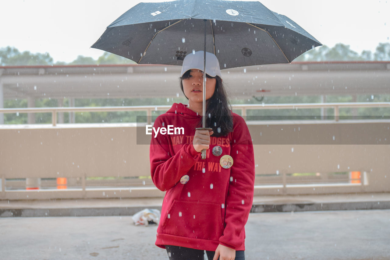 Young woman holding umbrella while standing on road during rainfall in city