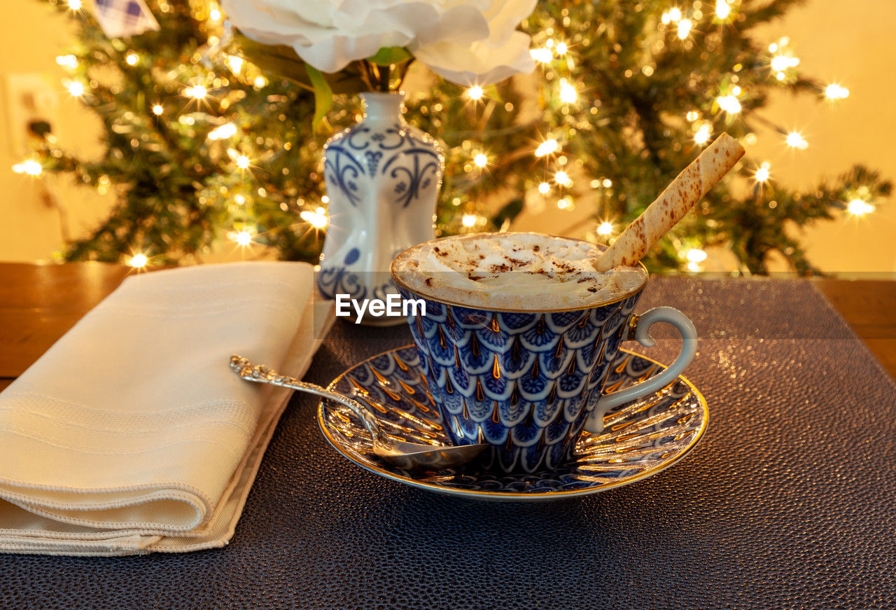 Cup of hot chocolate in a fancy bone china cup with gold and a silver spoon in front of a christmas