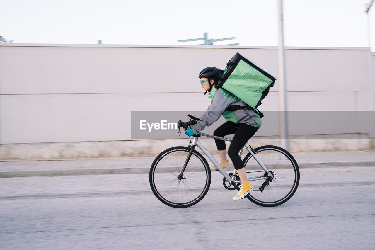 Side view of female courier with thermal bag riding bike on street road while delivering food in city, motion blur