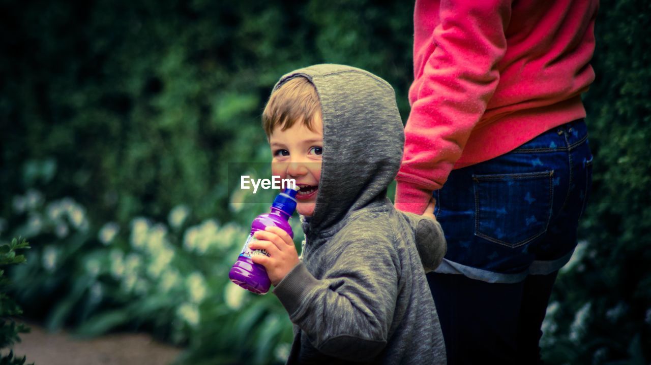 Portrait of happy boy holding water bottle while walking with mother against plants