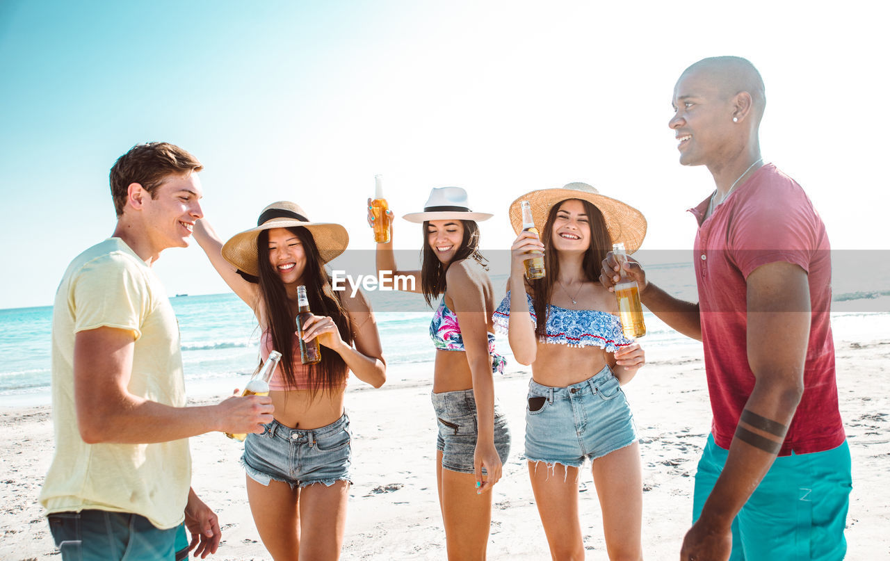 Friends holding drinks while standing at beach against sky