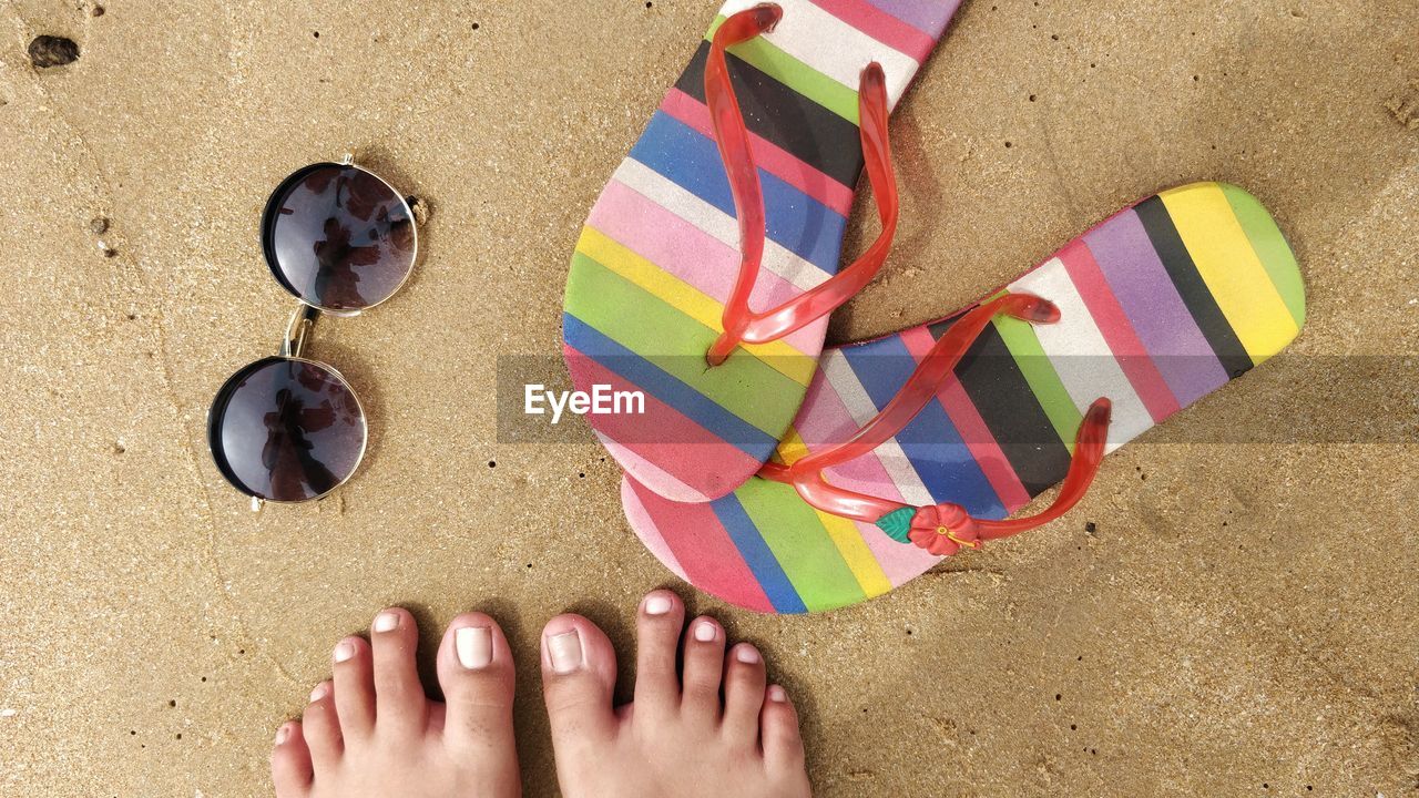 Low section of woman standing by flip-flops and sunglasses at beach