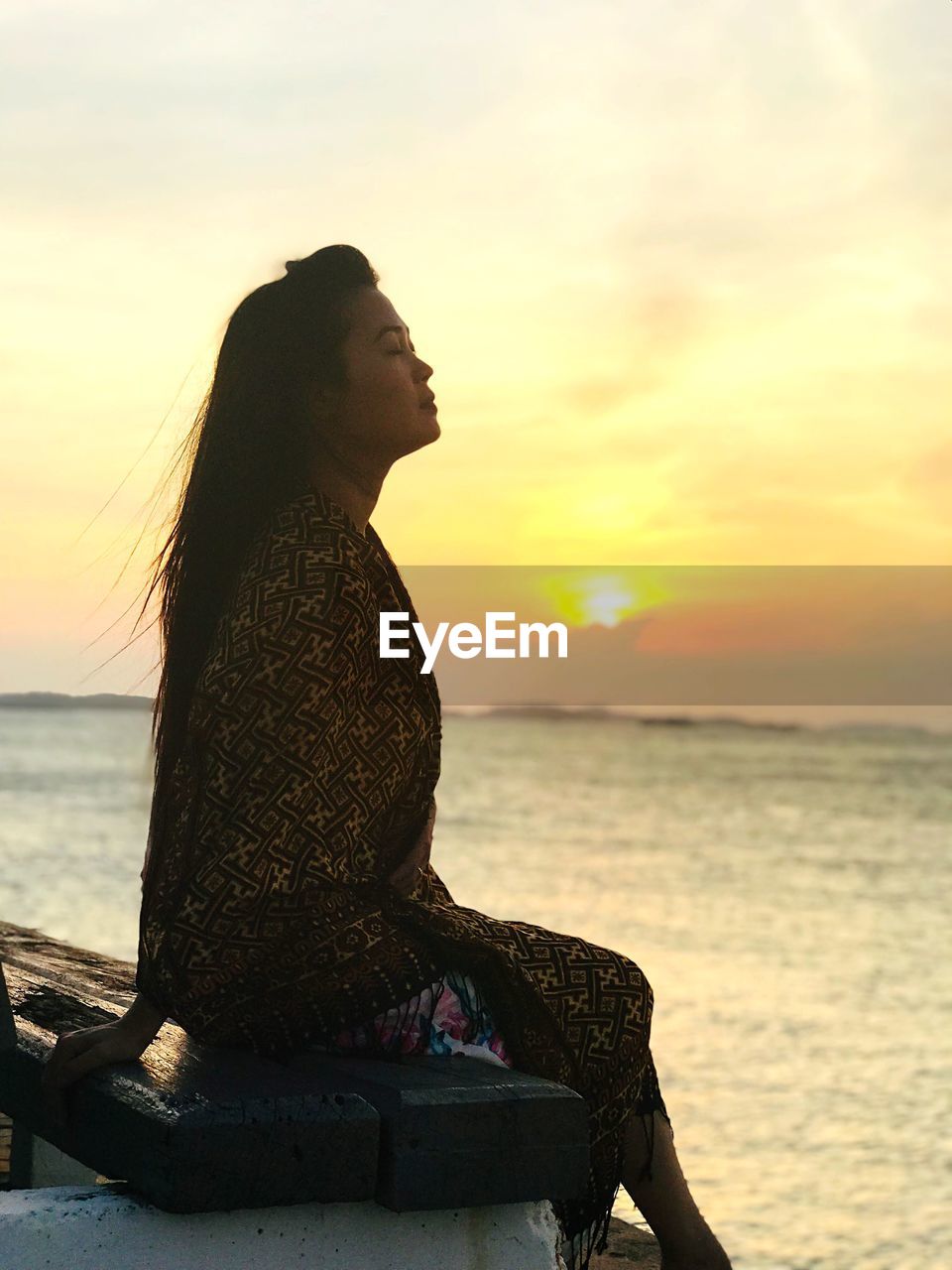 Woman sitting at beach against sky during sunset