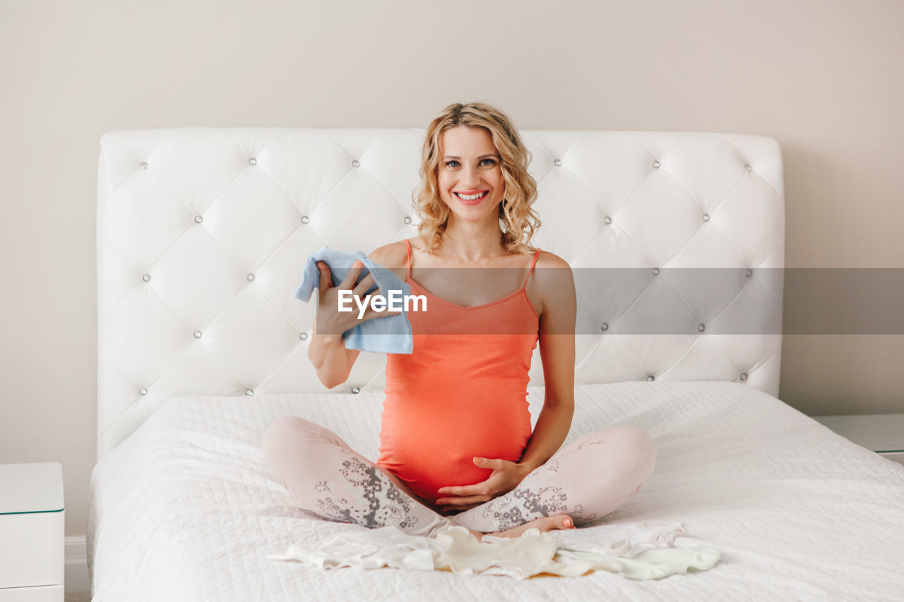Portrait of smiling pregnant woman holding baby clothing while sitting on bed at home