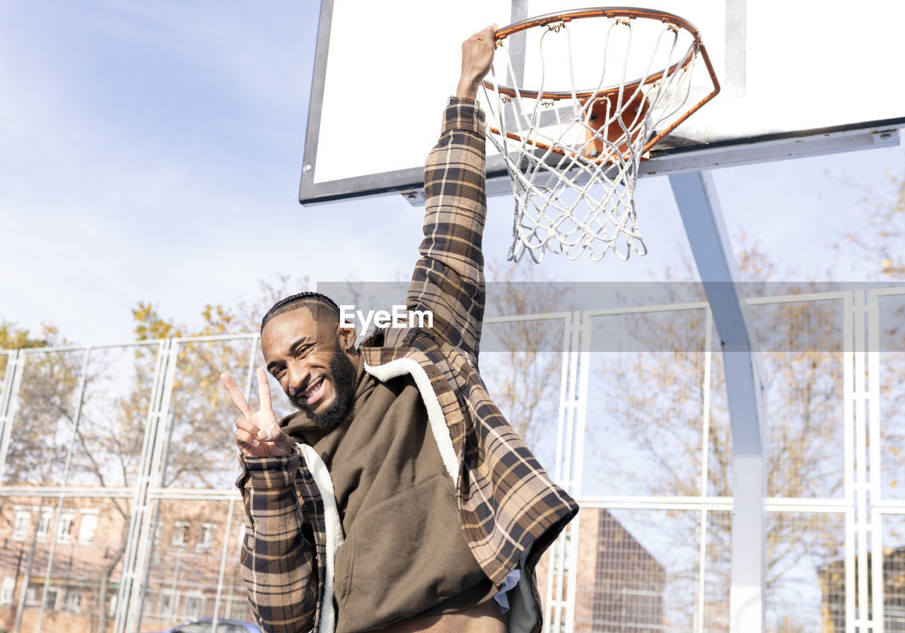 Happy young man gesturing peace while hanging on basketball hoop in court during sunny day
