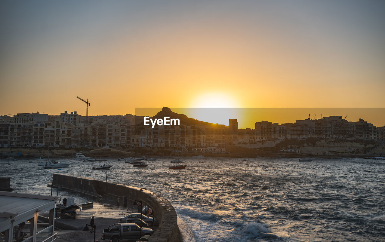 BUILDINGS BY SEA AGAINST SKY DURING SUNSET IN CITY