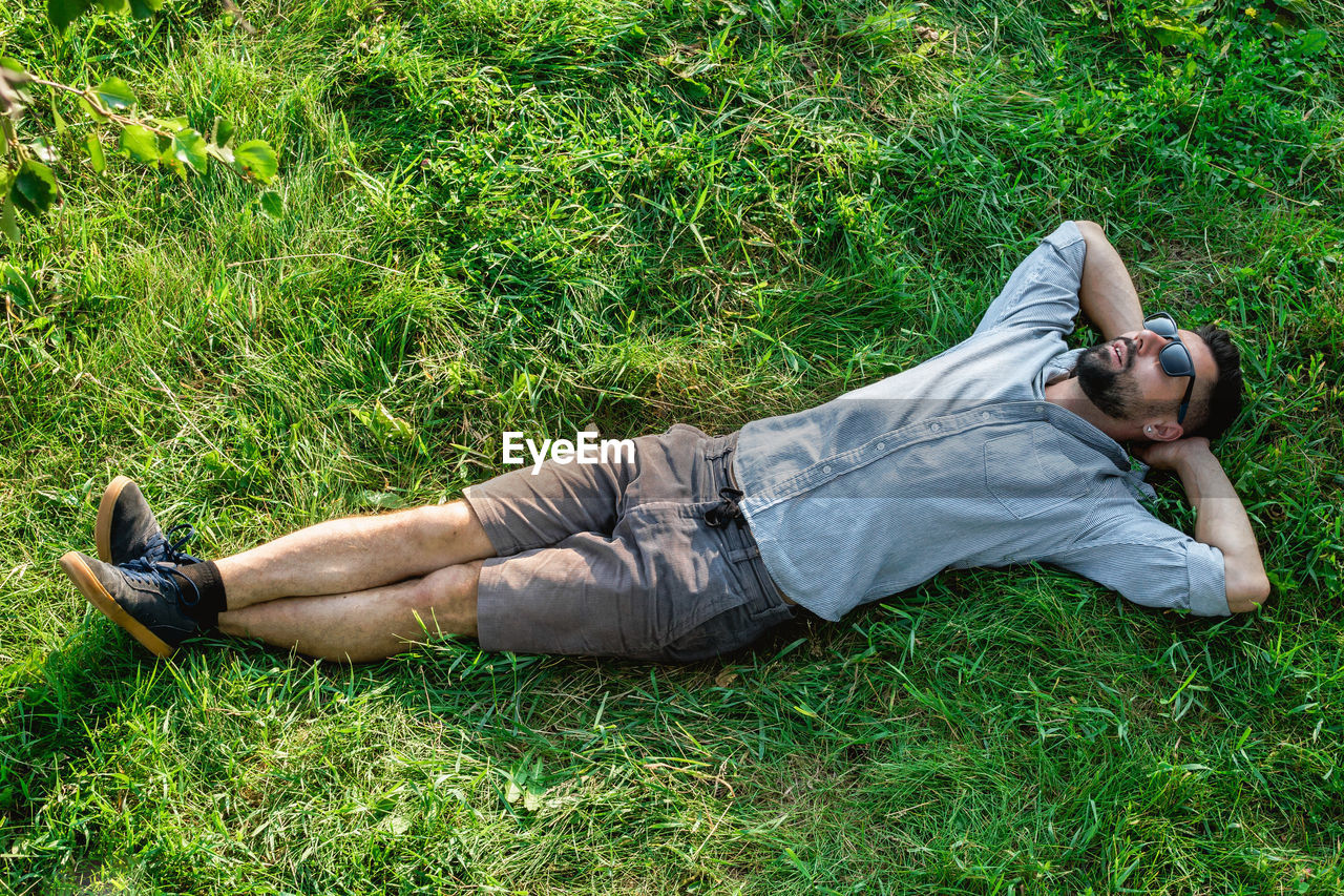 Young handsome sports european man in sunglasses is resting on a grass in summer park, top view.