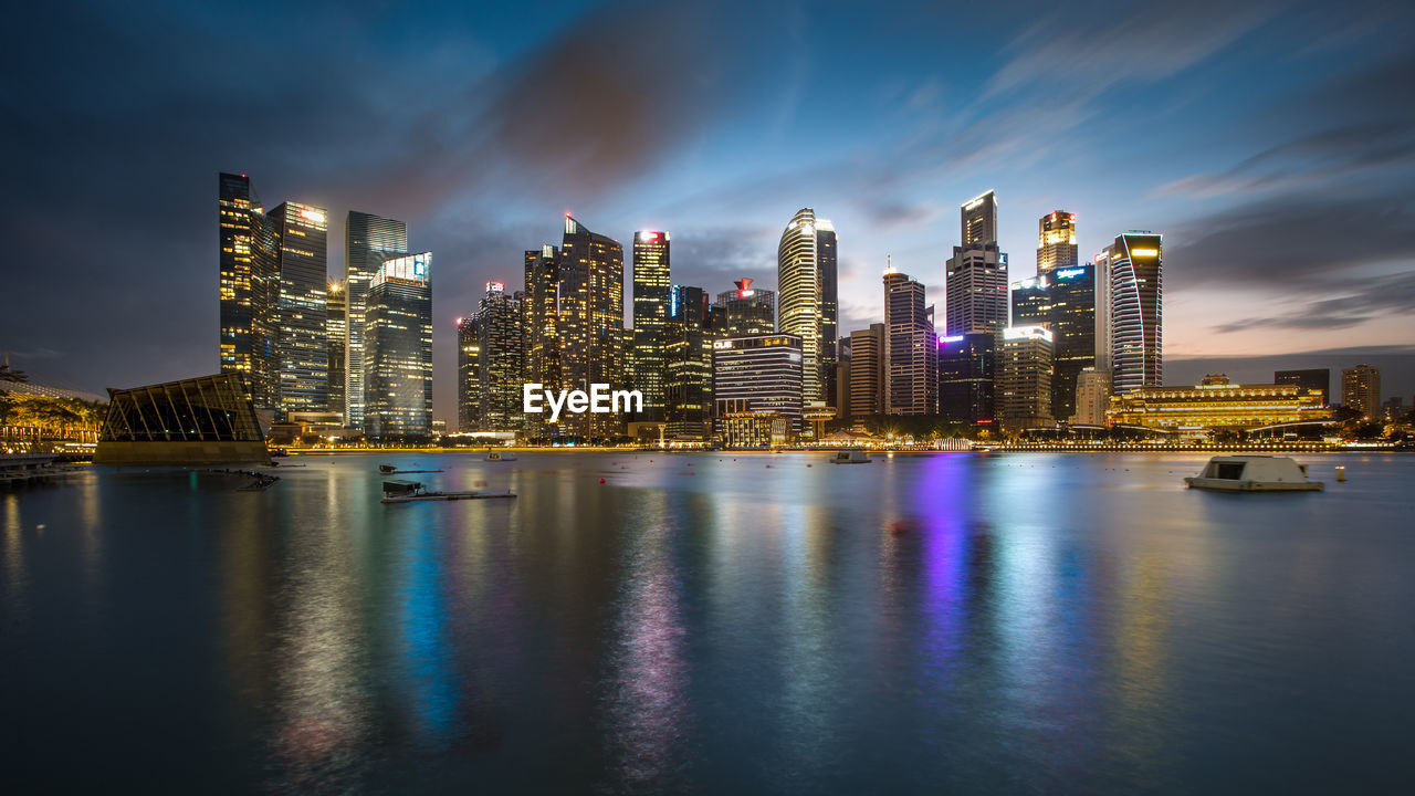 Illuminated buildings by river against sky in city