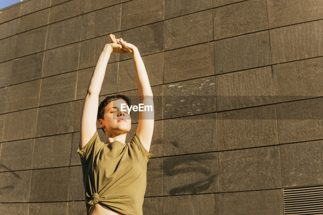 Woman stretching arms in front of wall on sunny day