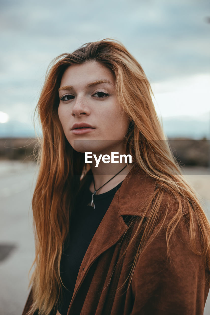 Confident young female with long ginger hair standing on street on cloudy day and looking at camera