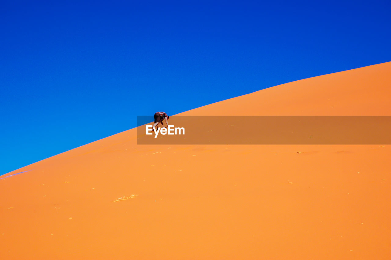 Man walking on sand dune against clear blue sky at desert