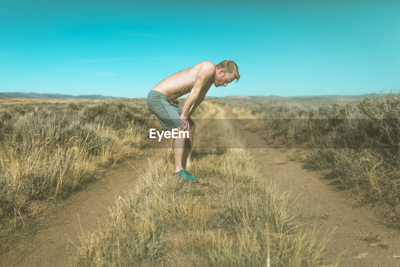 Side view of tired shirtless man relaxing on grassy field against clear blue sky