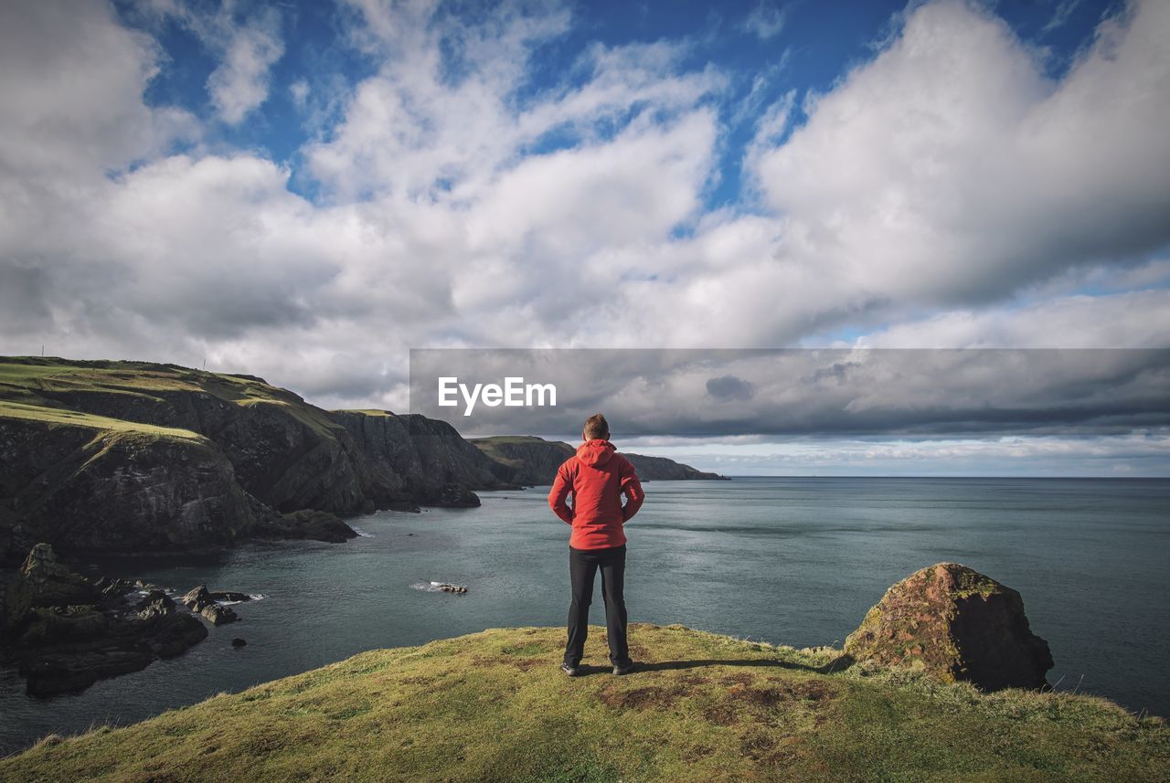 Rear view of man standing on cliff by sea against sky