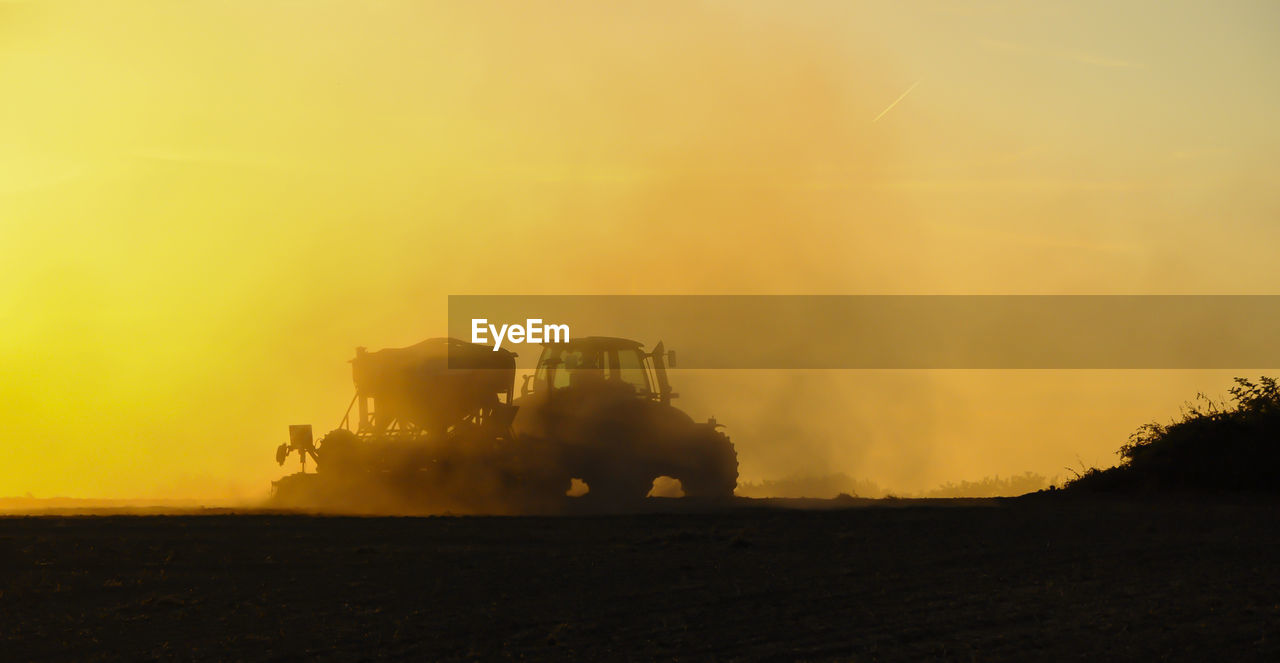 Silhouette of a tractor sowing seeds in a field in a cloud of dust against the background.