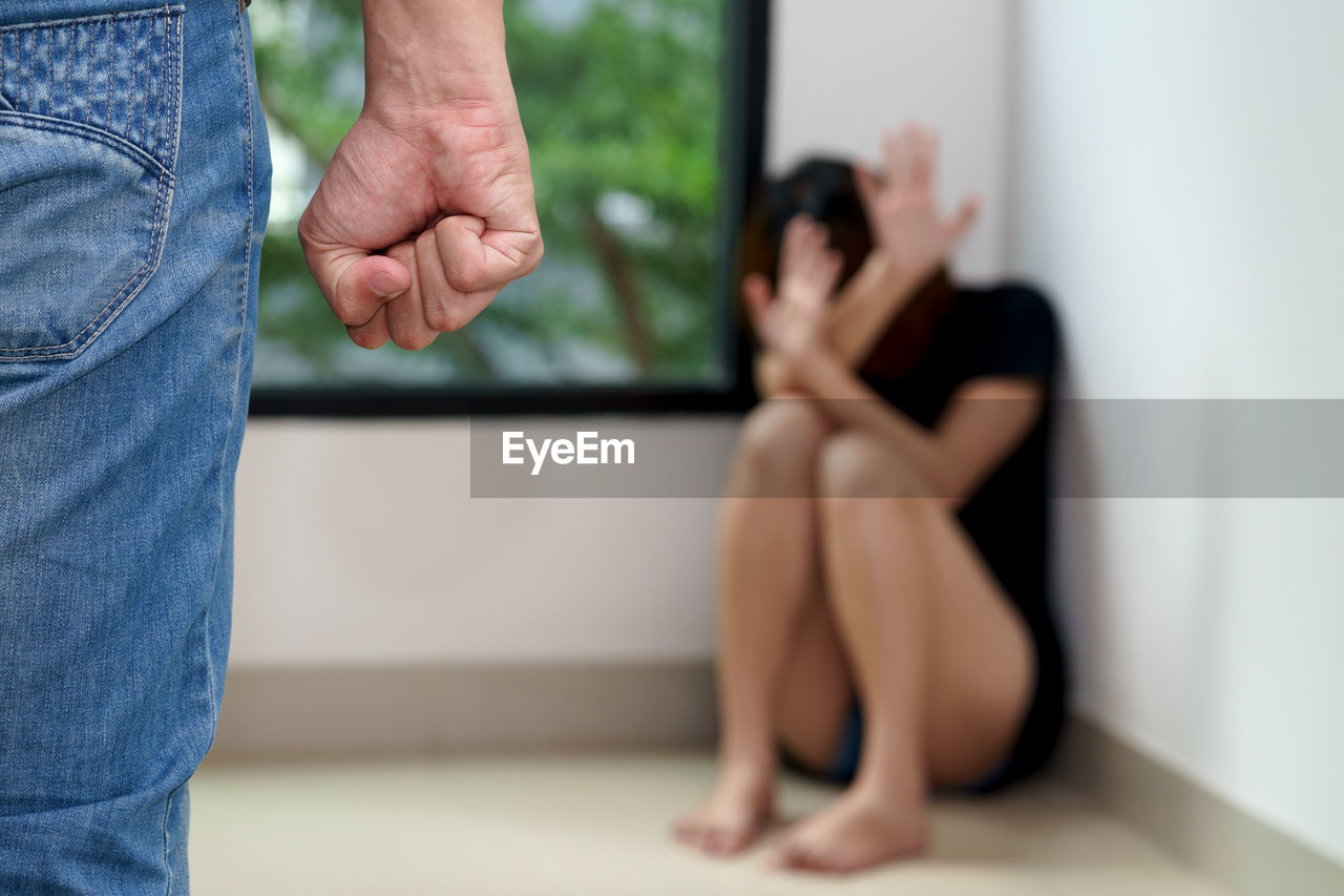 Midsection of man standing against scared woman sitting in corner