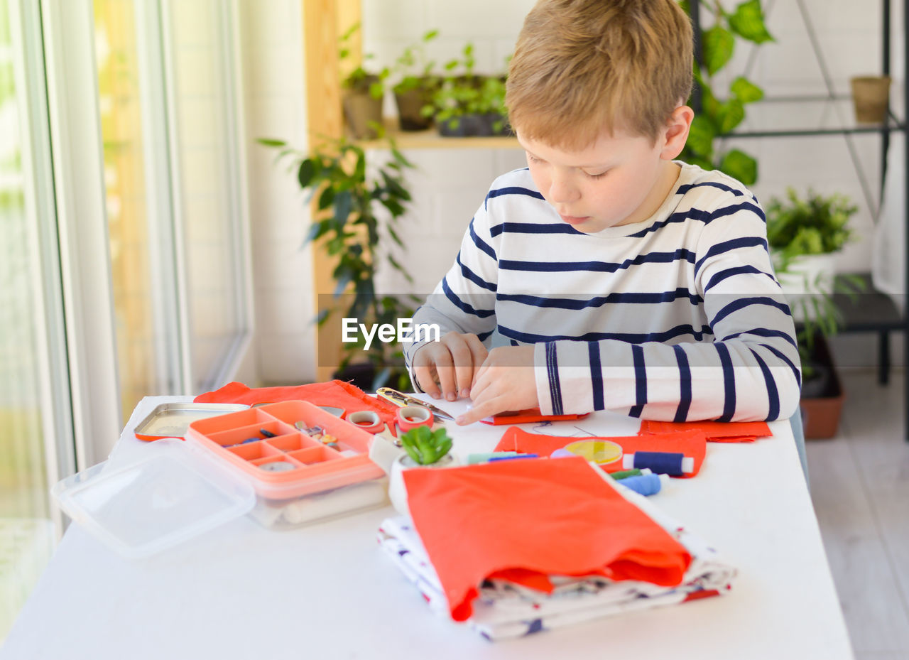 A caucasian boy of 9 years old is learning to sew with his hands homemade gift for mom for a holiday