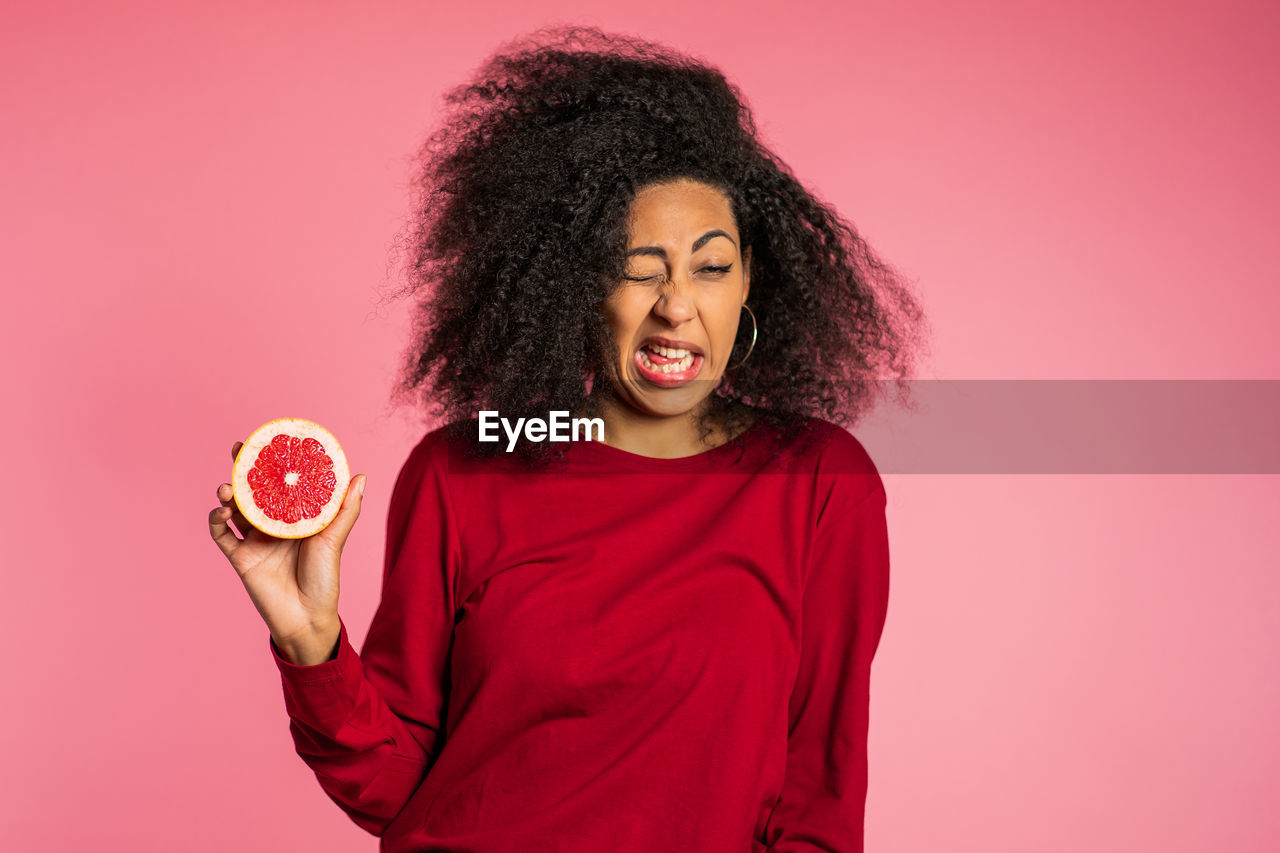 Young woman making face while holding grapefruit against pink background