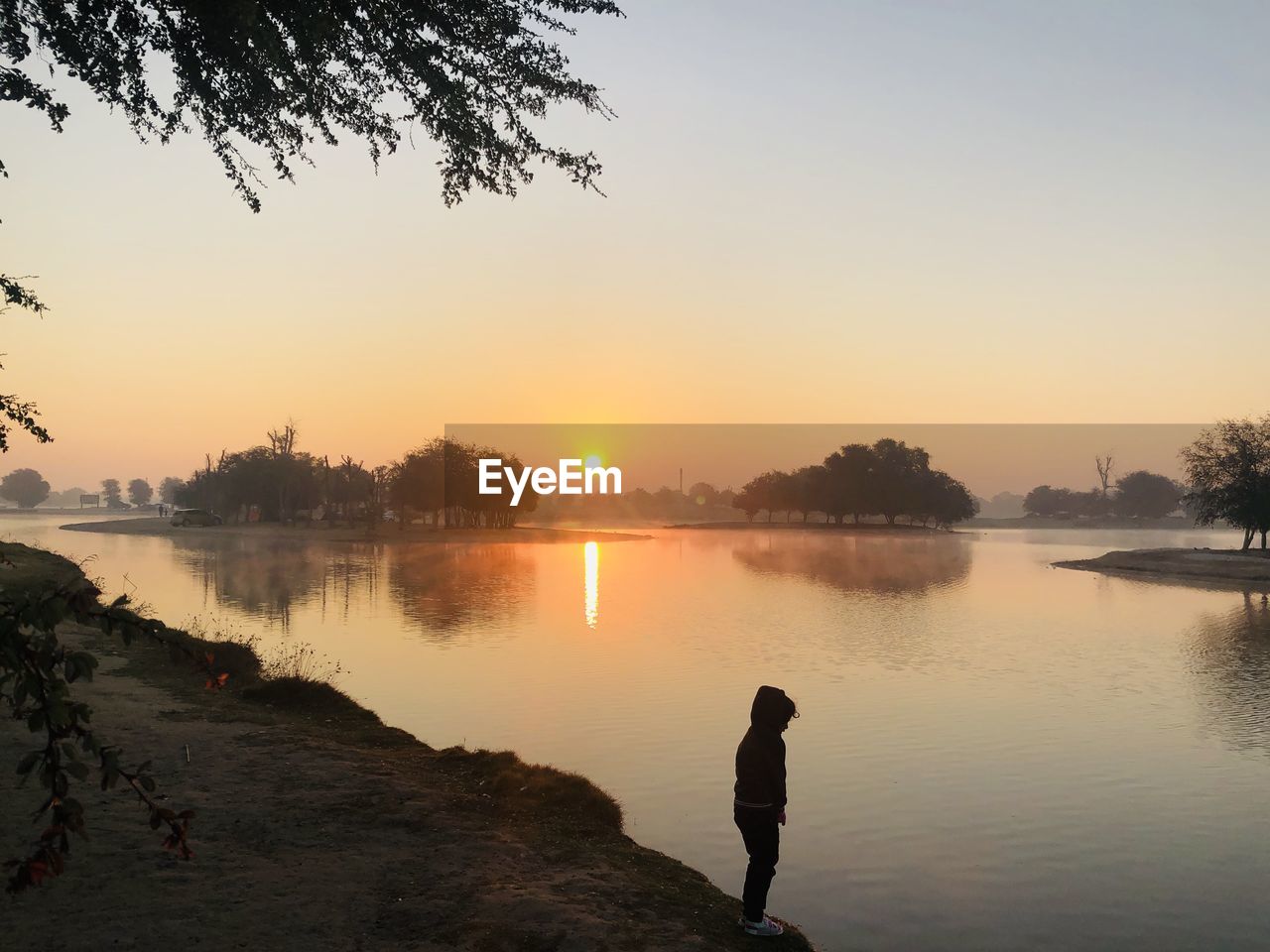 SILHOUETTE MAN BY LAKE AGAINST ORANGE SKY