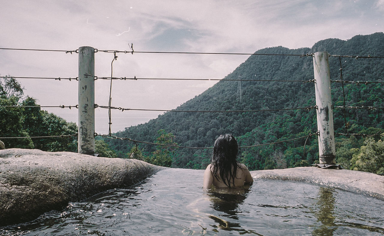 Rear view of shirtless person swimming in pond by tree mountains against sky