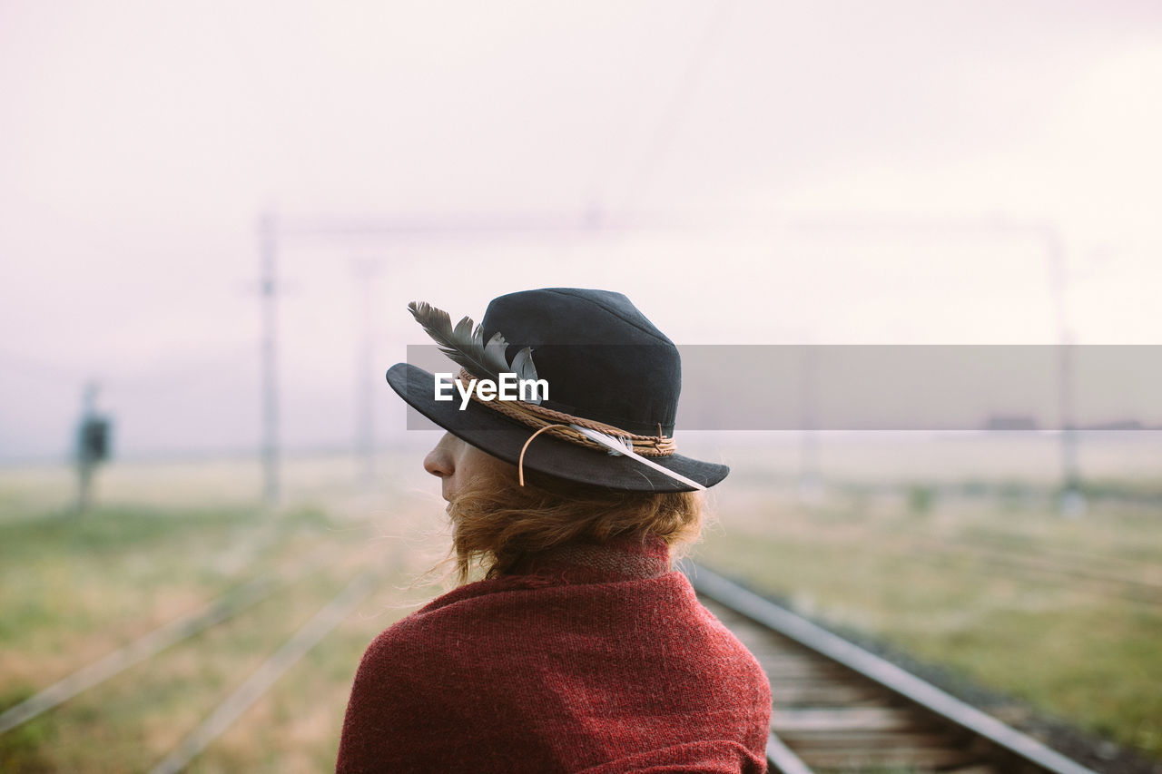 Rear view of woman wearing hat standing on railroad track against sky