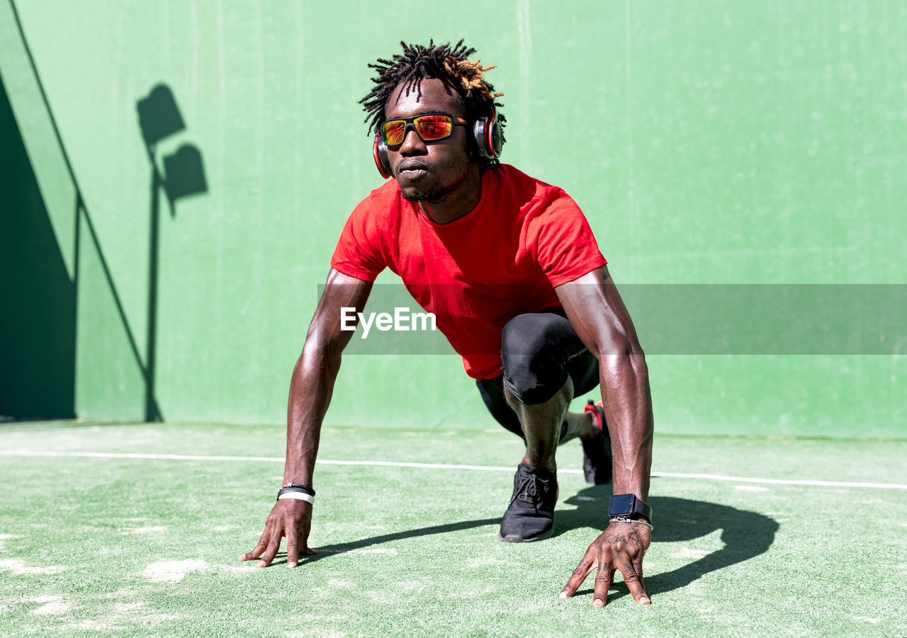 Determined black sportsman with dreadlocks stretching body while warming up before training and listening to music in headphones