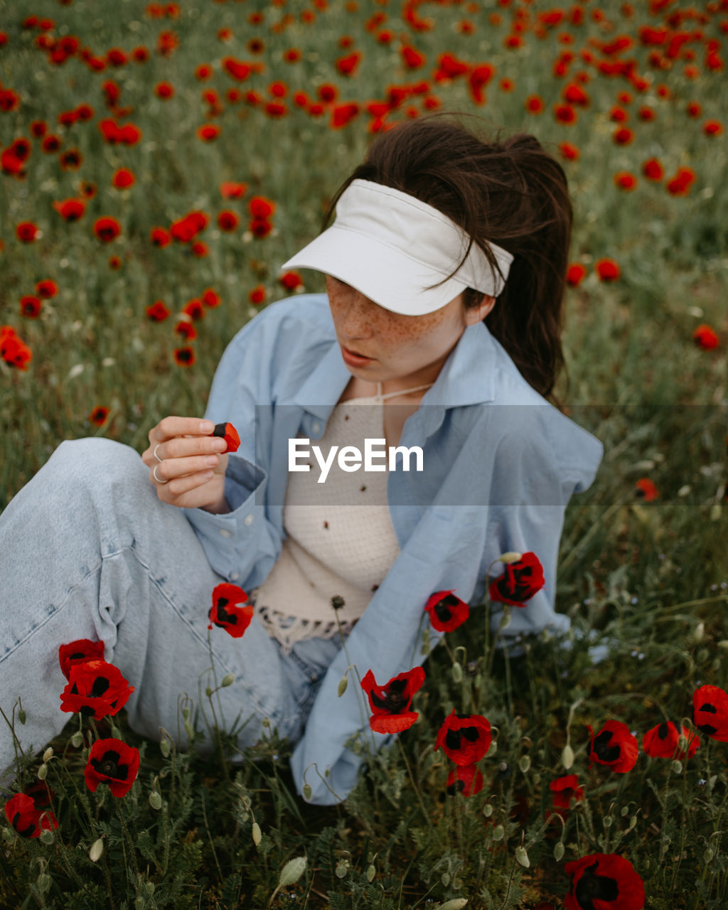 Attractive young woman sitting in the poppy field with flowers in blue shirt and jeans