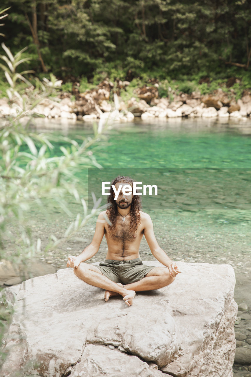 Shirtless young hipster meditating in lotus position on rock