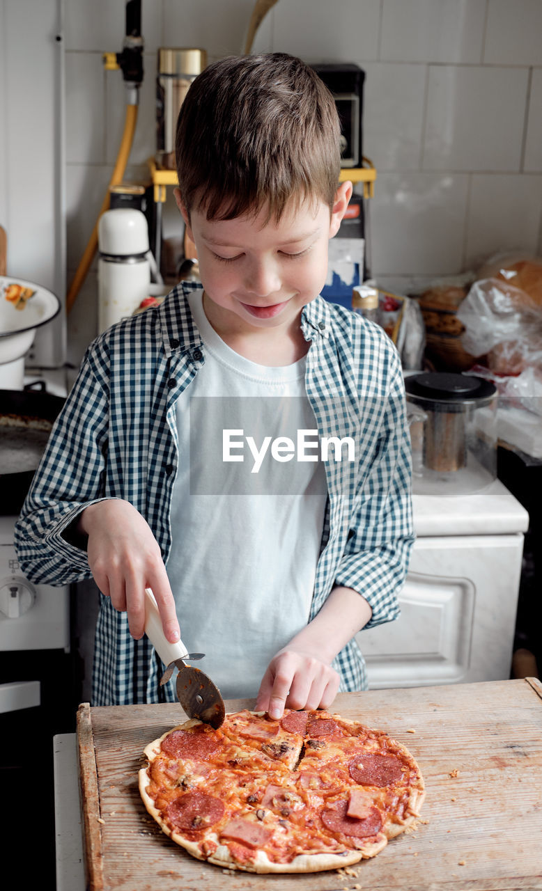 A caucasian boy in a shirt cuts pizza with salami with a round knife in the kitchen, on the table. 