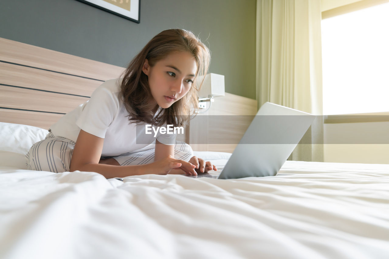 YOUNG WOMAN USING MOBILE PHONE WHILE SITTING ON BED