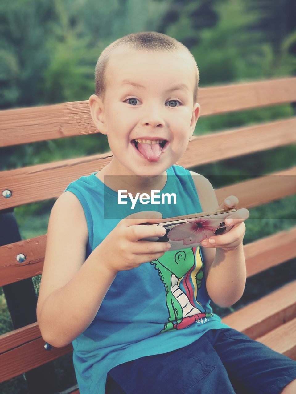 Portrait of boy with sticking out tongue holding mobile phone while sitting on bench