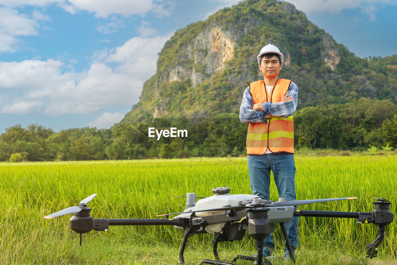 A farmer in a cap stands in a lush wheat field, directing a drone that is flying above the edge. 