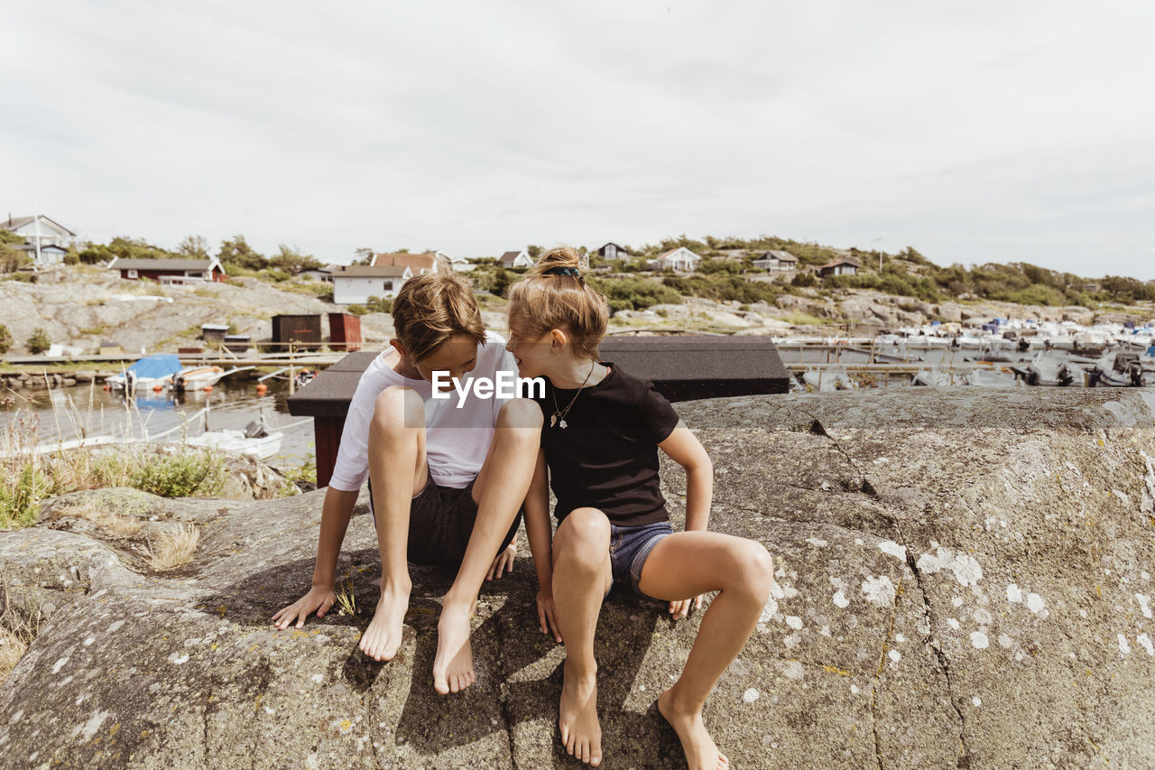 Happy siblings sitting on rock against sky during summer vacation