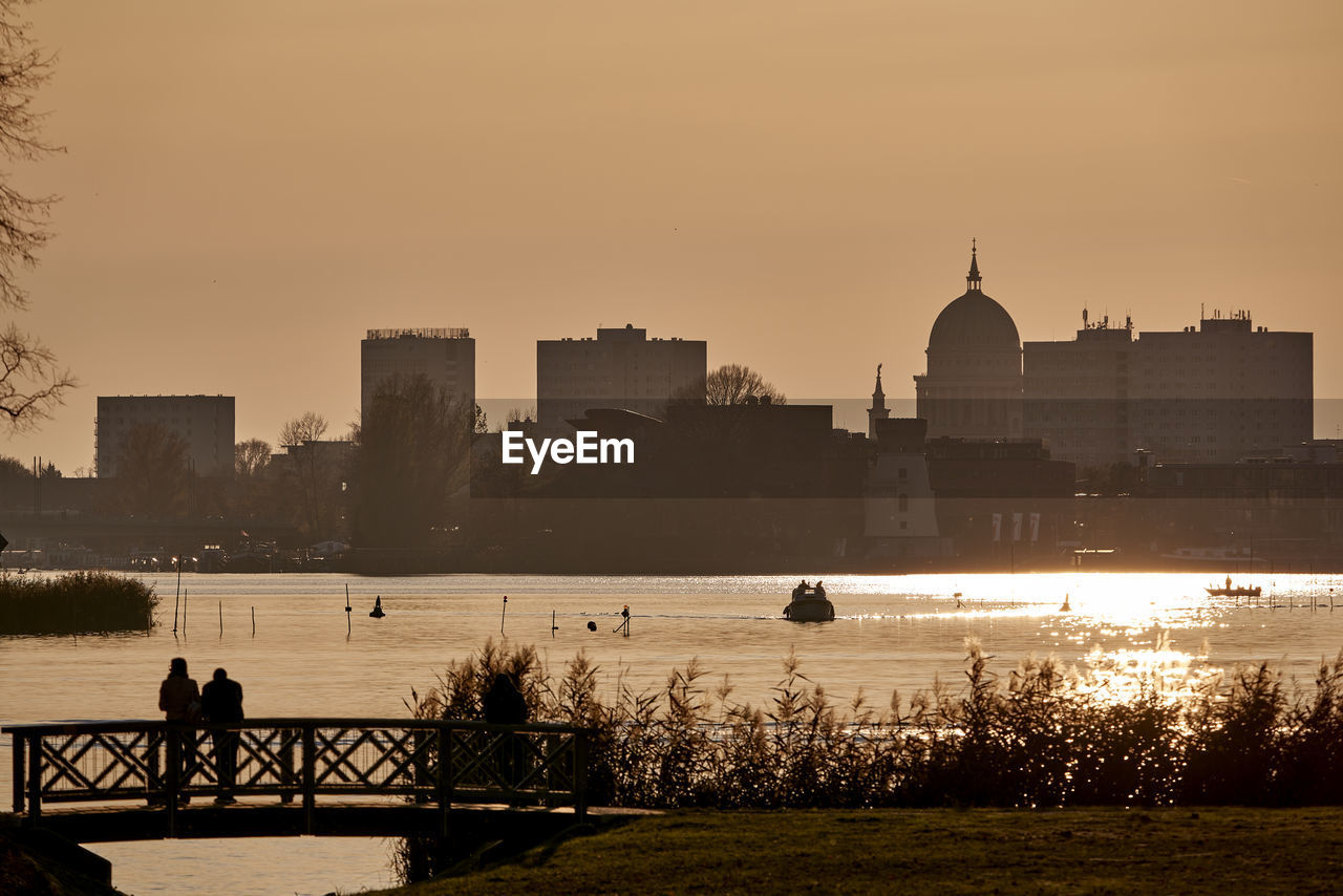 SILHOUETTE PEOPLE BY RIVER AGAINST BUILDINGS IN CITY AT SUNSET