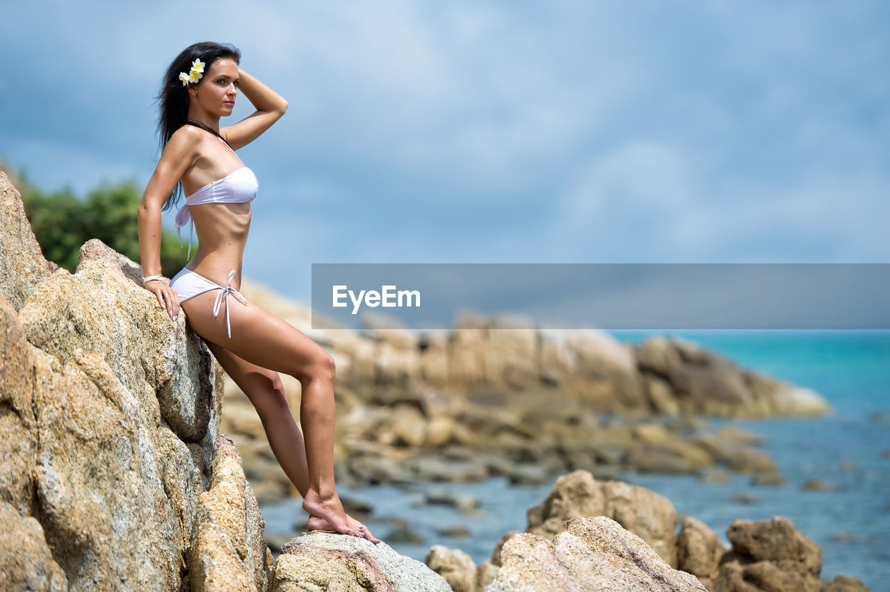 Full length of young woman in bikini leaning on rock by sea