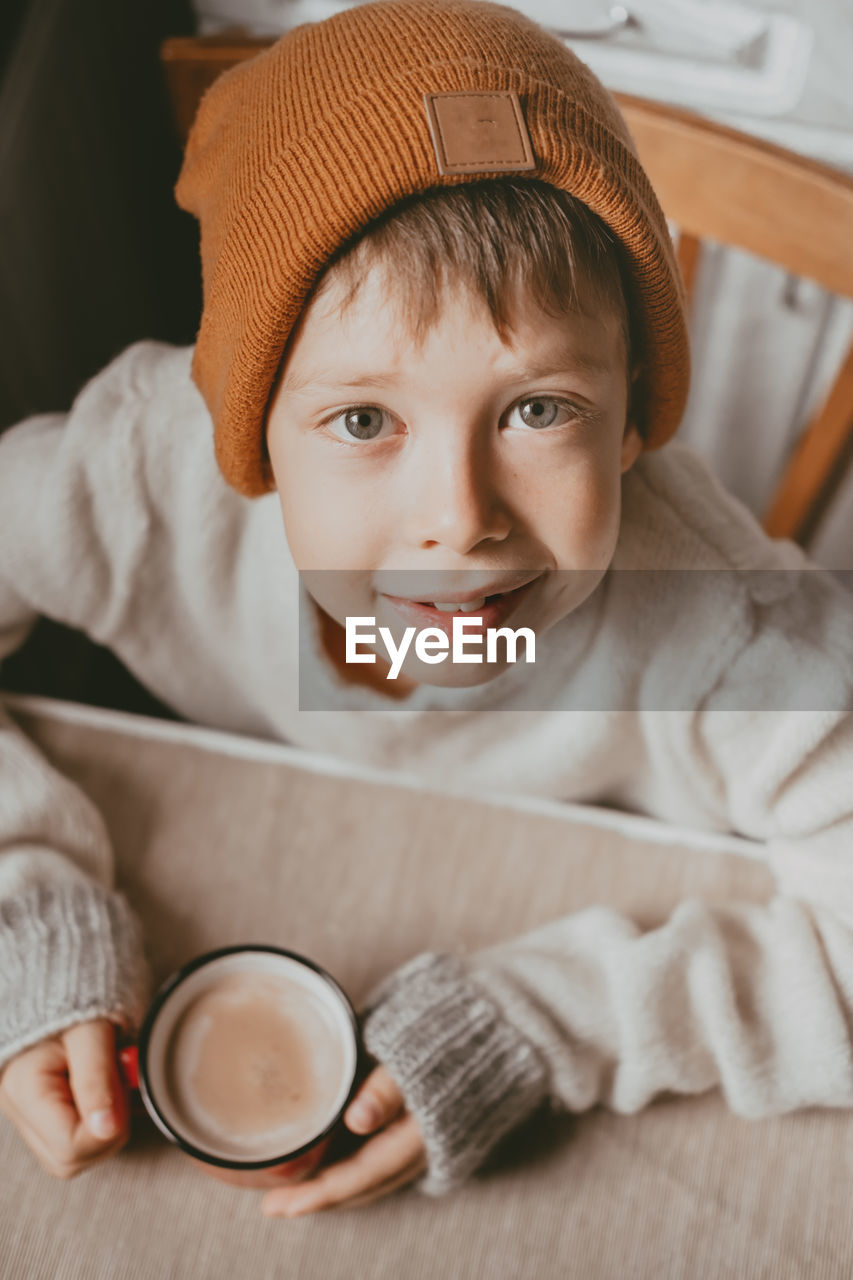 A boy in a sweater and a brown hat drinks cocoa from a red cup. a cozy photo with a mug in hand. 