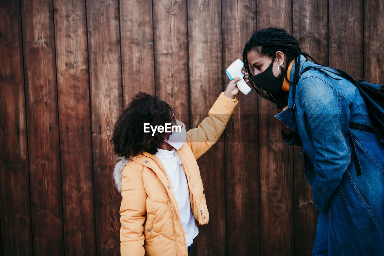 Girl checking woman temperature through infrared thermometer while standing against wooden wall