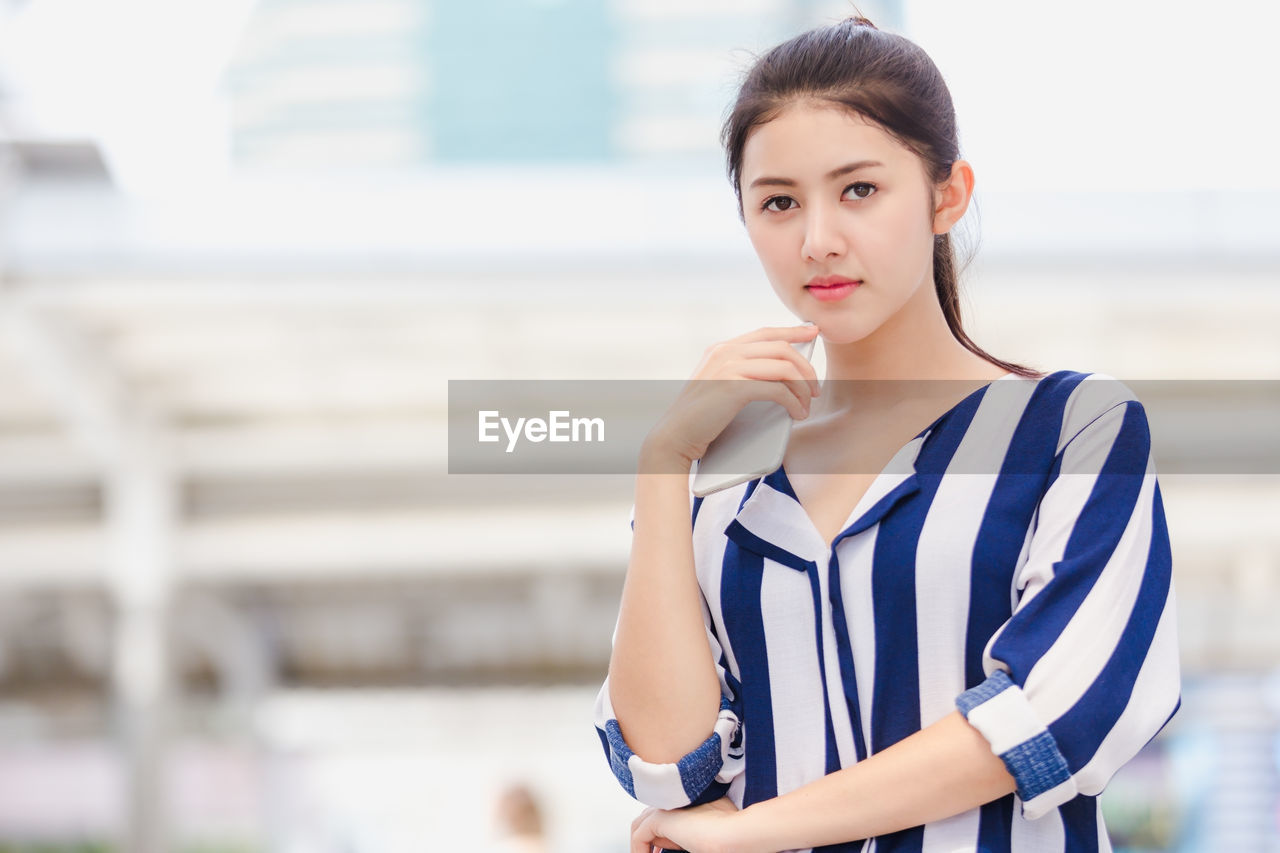 Portrait of young woman holding mobile phone while standing outdoors