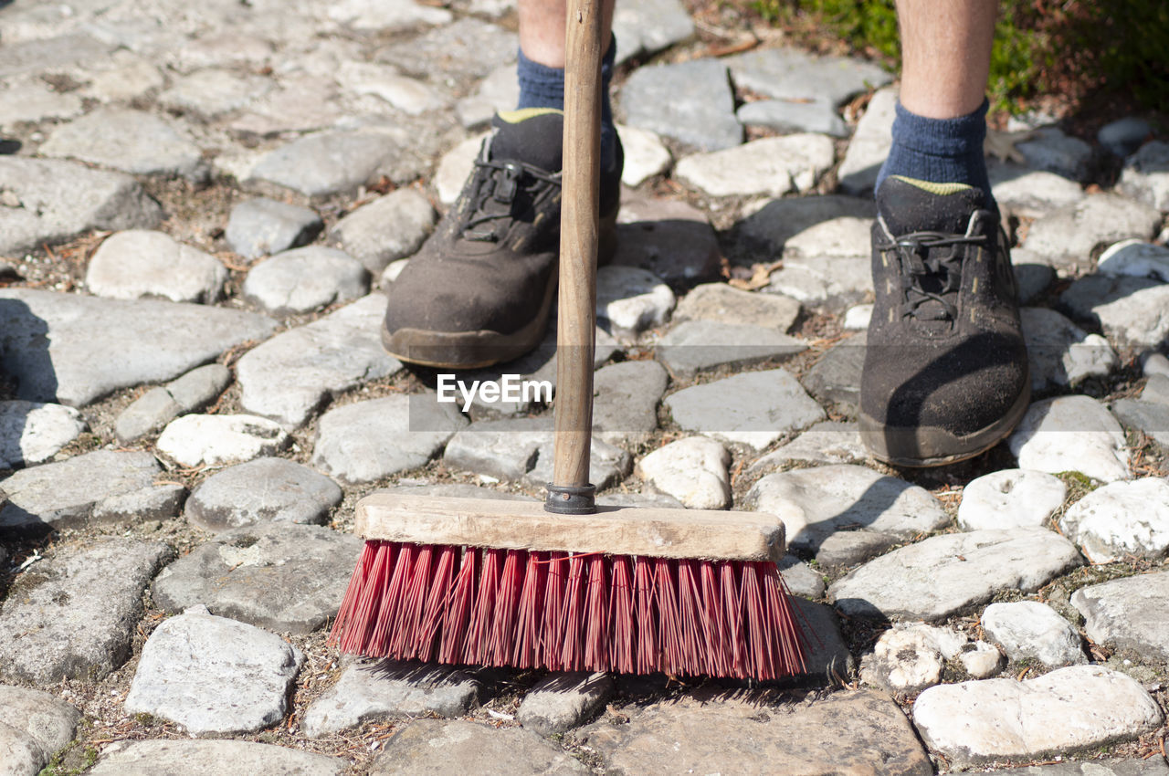 Gardener in dirty work clothes and boots holds garden tools, plastic brush broom