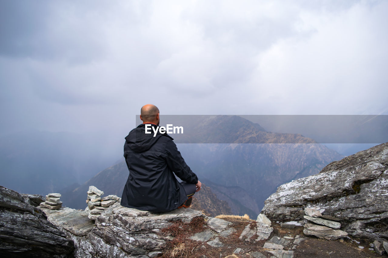 Rear view of mid adult man sitting on mountain against cloudy sky