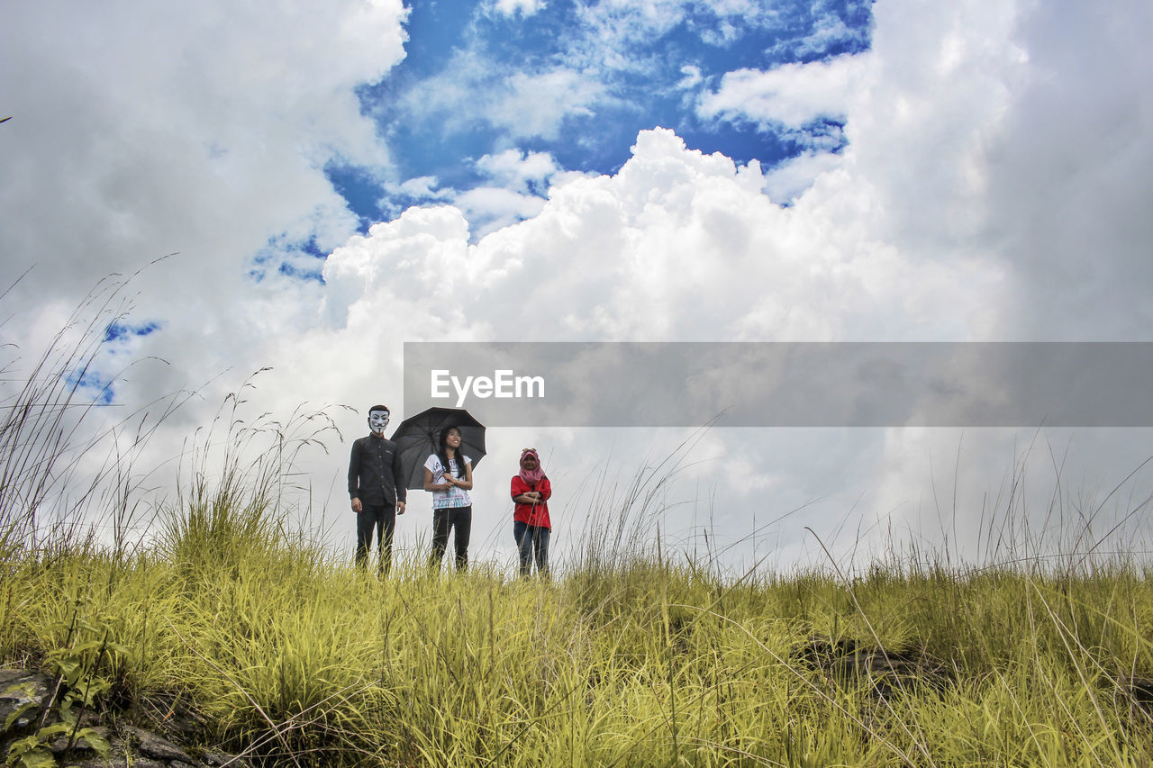 Low angle view of friends standing on grassy field against cloudy sky