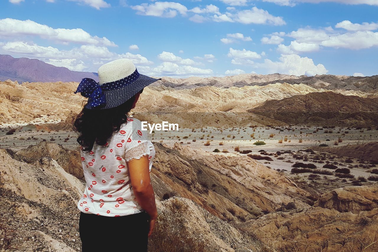 REAR VIEW OF WOMAN STANDING ON ROCK LOOKING AT MOUNTAIN RANGE