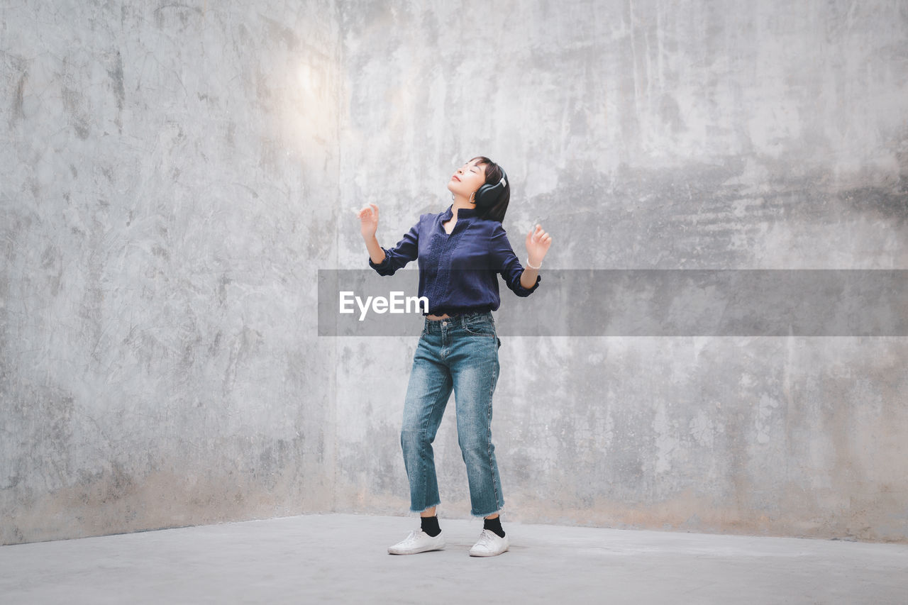 Full length of woman listening music while standing against wall