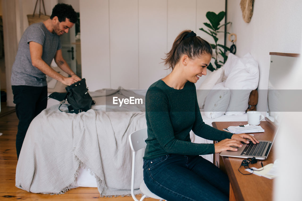 Smiling woman using laptop while boyfriend looking in purse at home