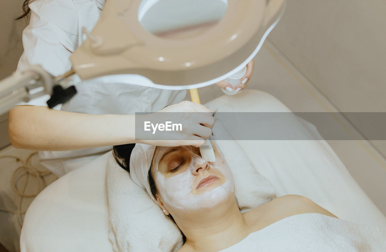 A girl cosmetologist applies a cream mask with a brush to a woman s face.