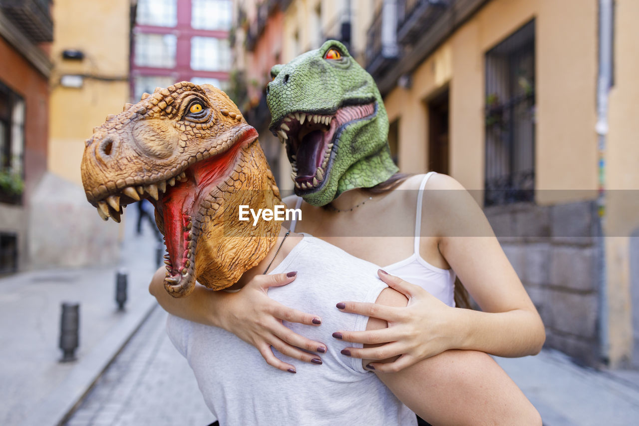 Male giving piggyback ride to female in dinosaur mask in city