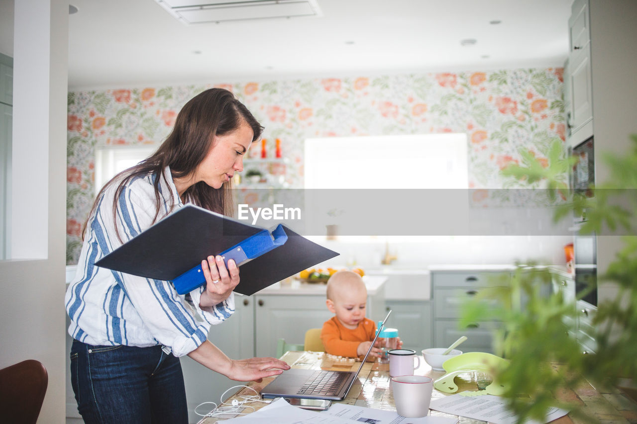 Multi-tasking mother telecommuting while daughter sitting at dining table in kitchen