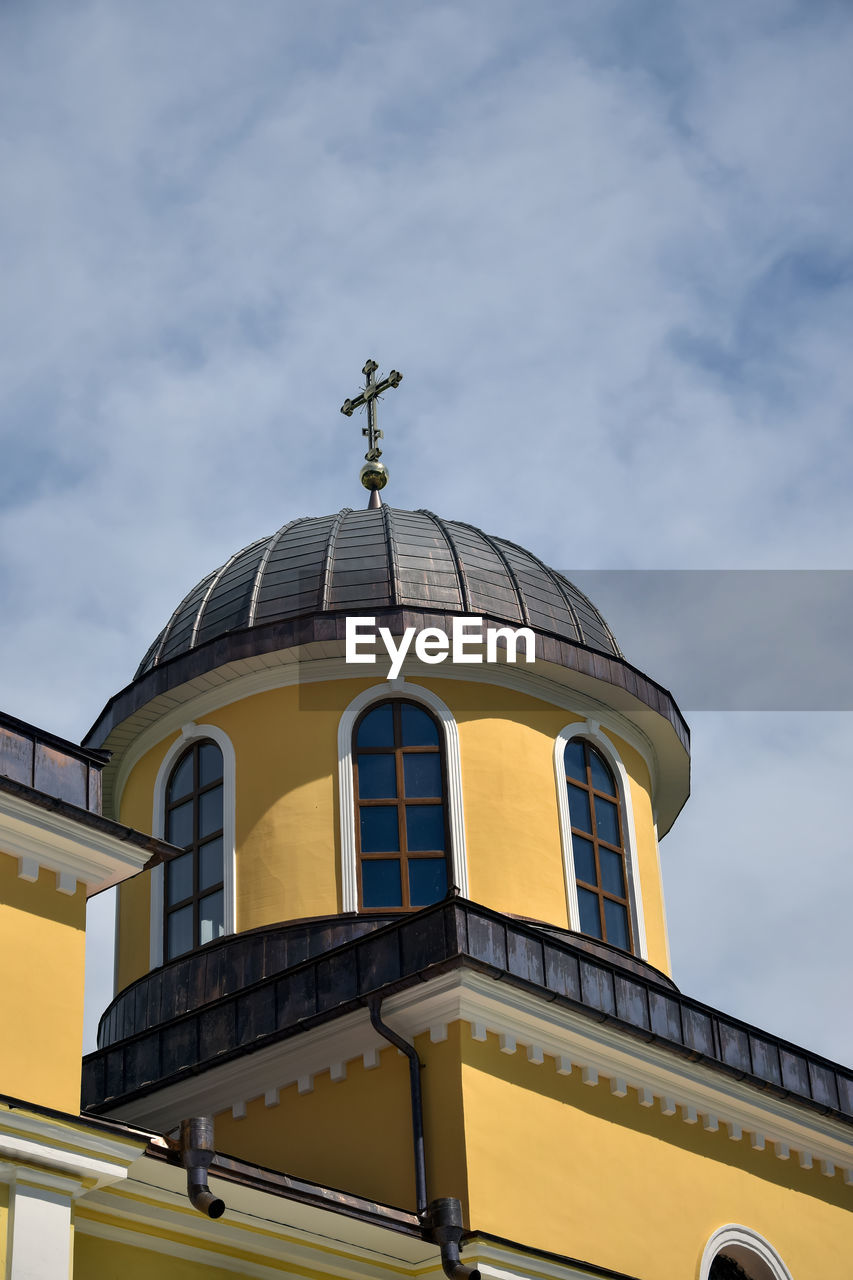 View from below of church tower with arched windows and metal dome with cross against cloudy sky.