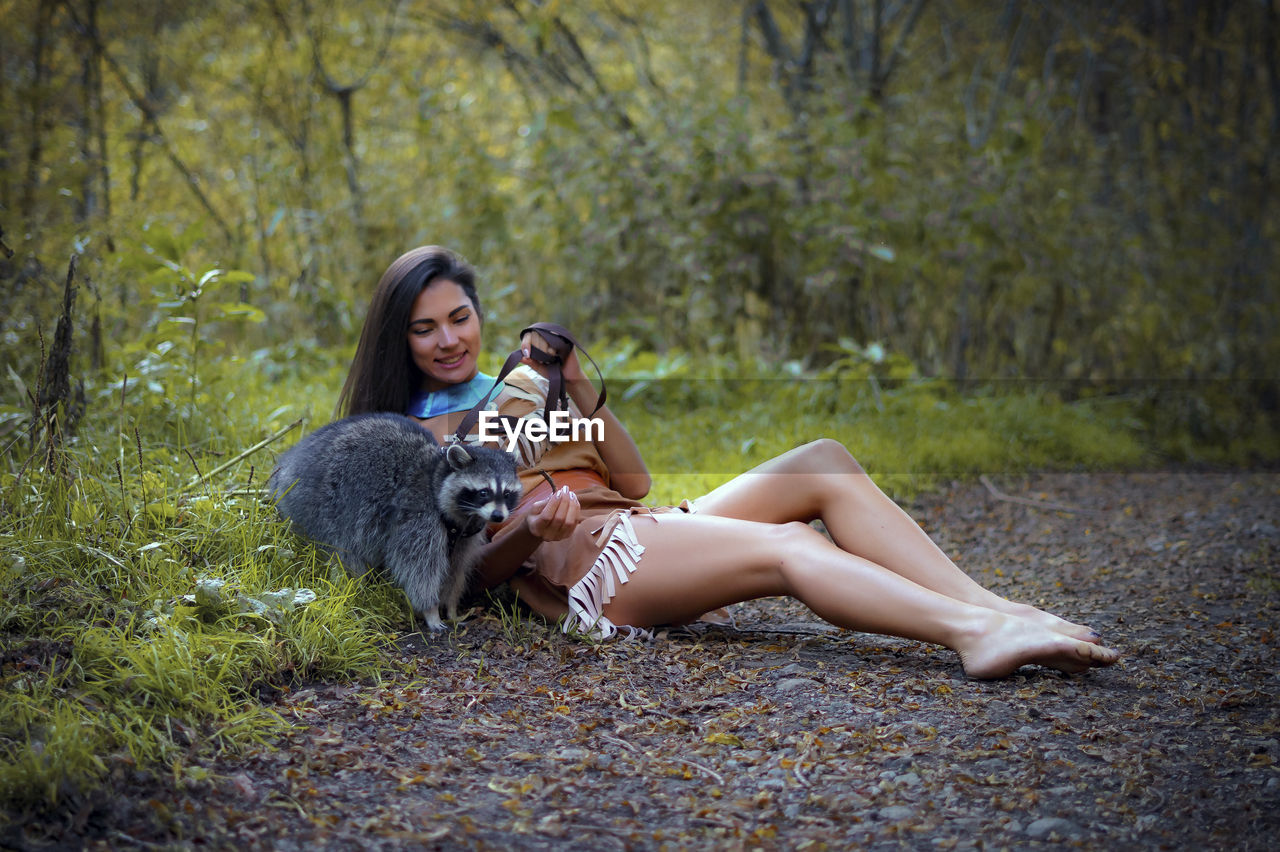 Smiling young woman in traditional clothing sitting by raccoon in forest