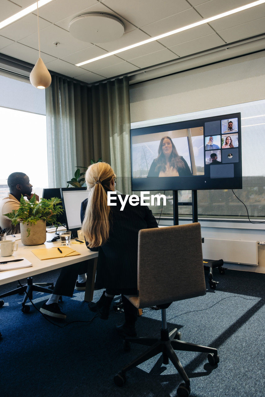 Male and female colleagues doing video call with businesswoman on tv in coworking office